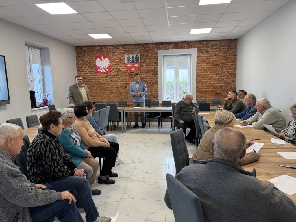 Mieszkańcy w sali z herbem Polski i herbem Gminy Oporów słuchają prowadzących spotkanie